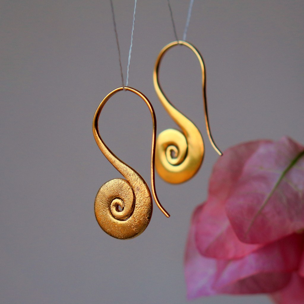 A pair of golden spiral earrings hanging against a soft background, with a hint of pink petals in the foreground.