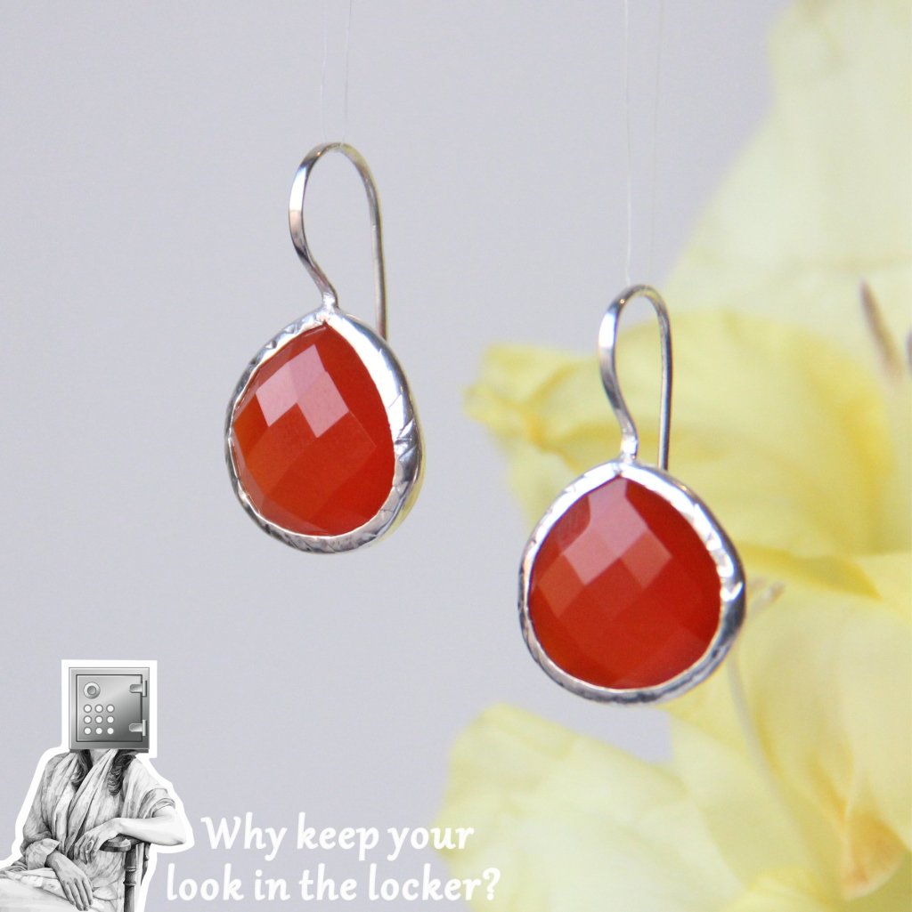 A close-up of handcrafted silver earrings featuring red faceted teardrop gemstones, set against a soft yellow background with a playful slogan.