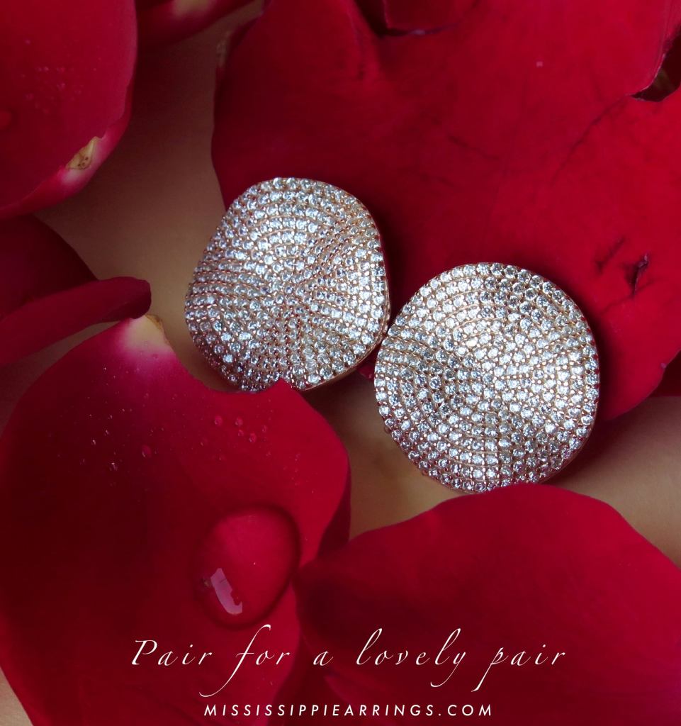 A pair of sparkling, round earrings resting on a bed of red rose petals, showcasing their elegant design.