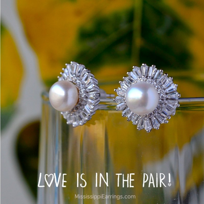 A close-up of elegant pearl earrings with a shiny silver setting, featuring a round pearl surrounded by a decorative halo of clear gemstones. The background showcases green and yellow foliage.